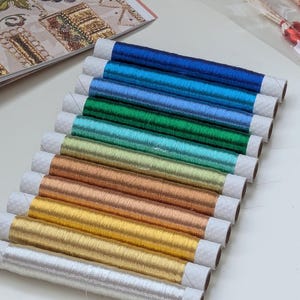 Op de afbeelding: Een collectie van twaalf klossen metallic borduurgaren gerangschikt in een diagonale rij. De klossen vertonen een kleurverloop met onder andere koningsblauw, turkoois, groen, blauwgroen, limoengroen, goud, koper, brons, geelgoud en zilver.