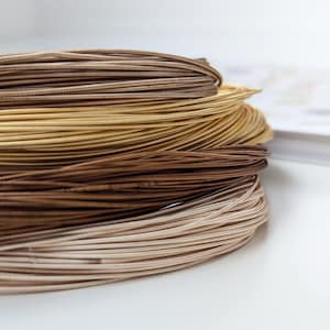 Puede incluir: Una colección de alambre metálico enrollado en varios tonos de marrón, dorado y beige. El alambre presenta una textura estriada o enrollada distintiva a lo largo de su longitud, dispuesta en capas circulares apiladas que crean una forma cilíndrica.
