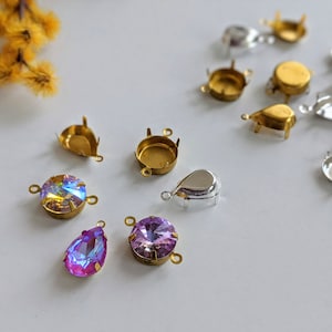 Könnte beinhalten: Sortiment aus goldenen und silbernen Metallfassungen für Strasssteine und Kristalle. Die Fassungen haben Ösen zum Anbringen an Schmuck. Es gibt zwei schillernde Strasssteine, einen runden und einen tropfenförmigen.