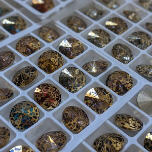 May include: A close-up of a white tray filled with round, iridescent rhinestones. The rhinestones have a leopard print pattern and are arranged in rows.