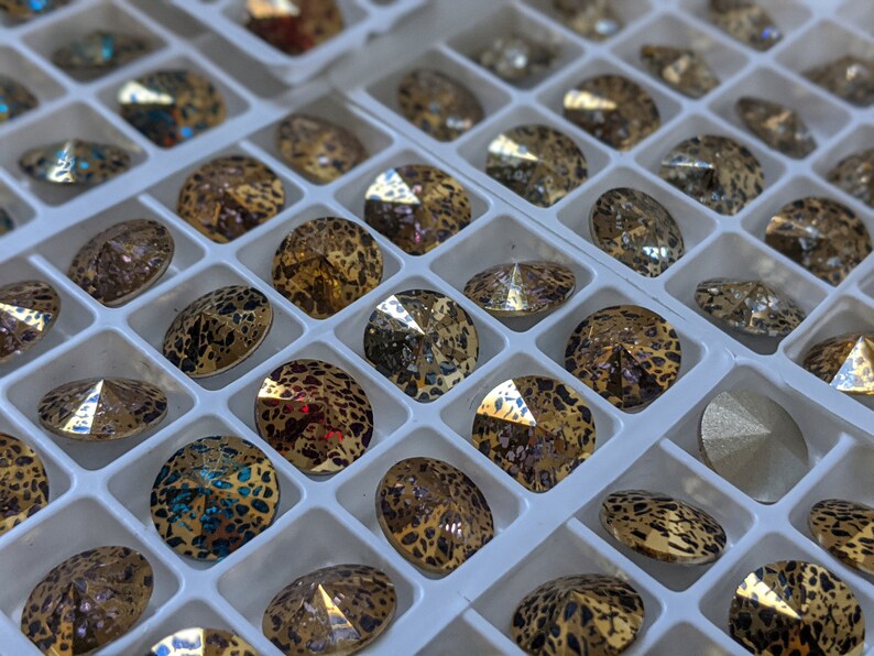 May include: A close-up of a white tray filled with round, iridescent rhinestones. The rhinestones have a leopard print pattern and are arranged in rows.