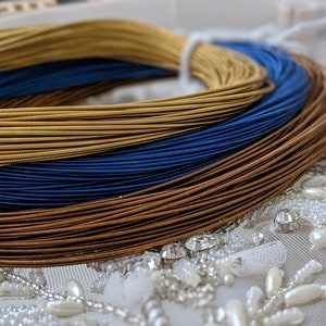 May include: Three spools of metallic thread, one gold, one blue, and one brown, are stacked on top of each other. The spools are on a white fabric with silver and white beads.