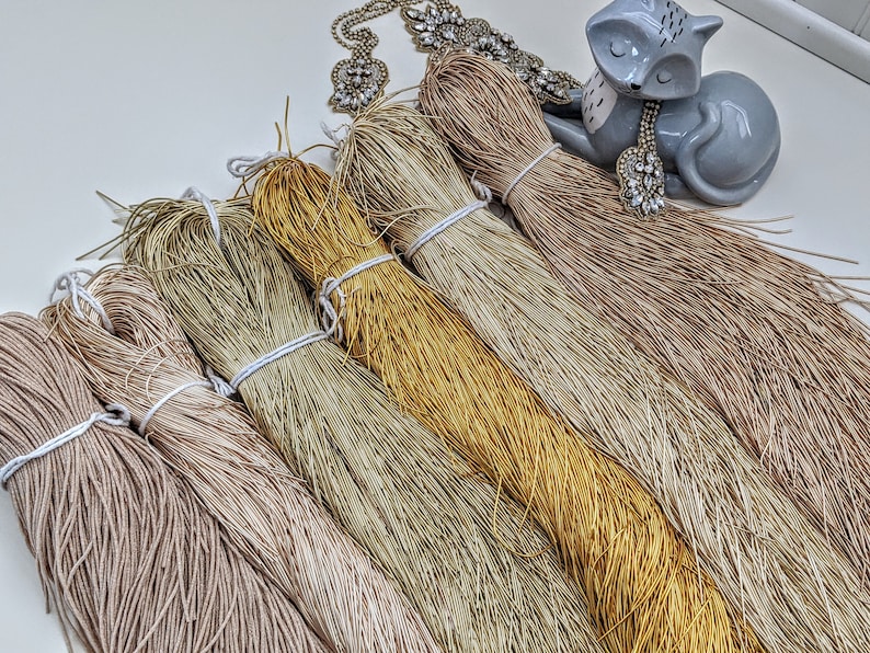 May include: A collection of six spools of natural fiber cord in various shades of beige, brown, and gold. The cords are arranged in a fan shape, with the ends of the cords extending out from the center.