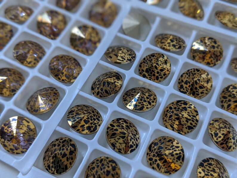 May include: A close-up of a tray filled with small, round, gold-toned rhinestones. The rhinestones have a black leopard print design.