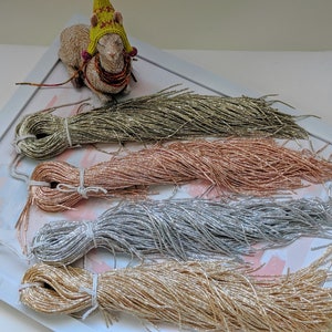 May include: Four spools of metallic thread in gold, rose gold, silver, and a green-gold color. The spools are arranged in a row on a white surface with a white frame around them.