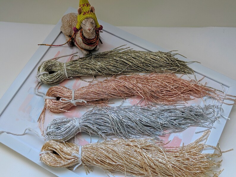 May include: Four spools of metallic thread in gold, rose gold, silver, and a green-gold color. The spools are arranged in a row on a white surface with a white frame around them.