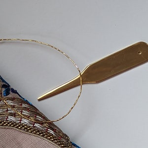 May include: A gold-colored embroidery needle threader tool with a small hole at the top. The tool is lying on a white surface with a piece of gold thread wrapped around it.