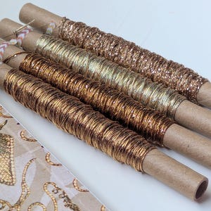 May include: Four cardboard tubes wrapped with metallic thread in shades of copper, bronze, gold, and rose gold. Each spool is secured with a small piece of patterned adhesive tape at the end, displaying a shiny, reflective texture on the thread.