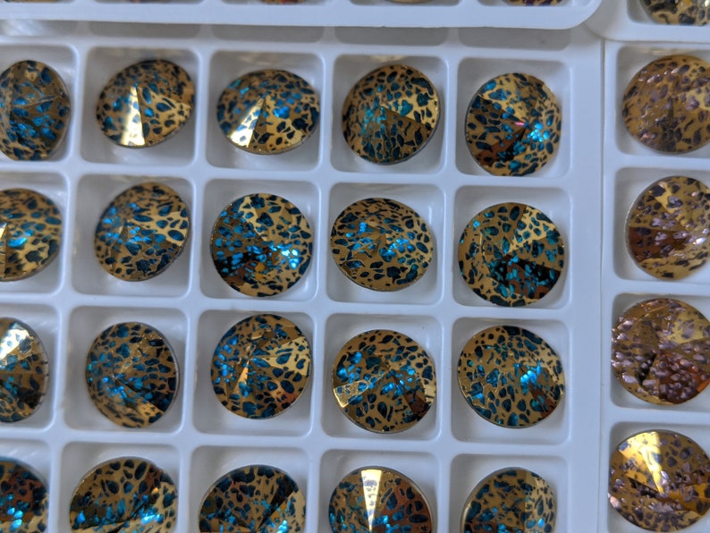 May include: A close-up of a tray of round, iridescent blue and gold rhinestones. The rhinestones are arranged in a grid pattern and have a textured, speckled surface.