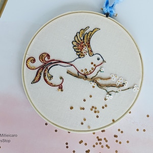May include: Embroidered hoop art featuring a white bird with gold and red accents perched on a branch with white blossoms. The background is a light pink with gold sequins scattered around the hoop.