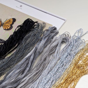 May include: A close-up of embroidery floss in various colors, including black, gray, silver, gold, and white. The floss is arranged in a fan shape, with the black floss at the top and the white floss at the bottom. The floss is used for embroidery projects.