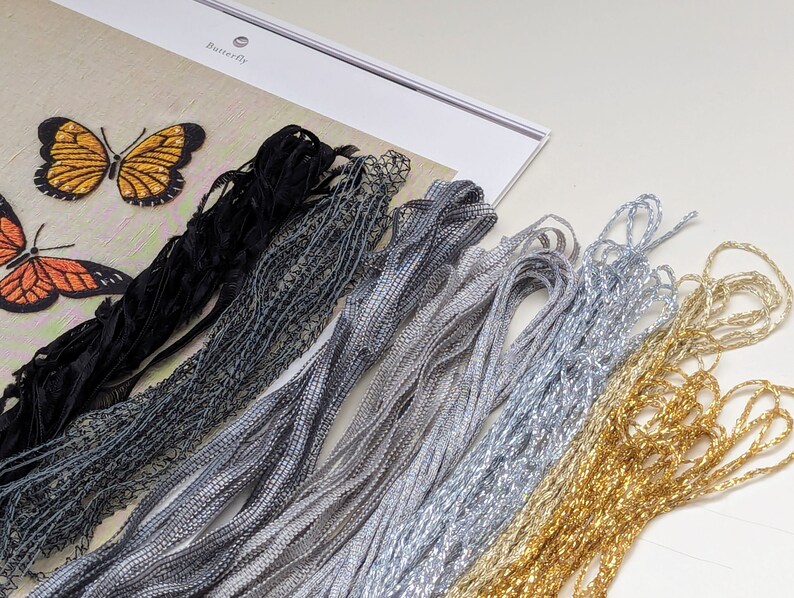 May include: A close-up of embroidery floss in various colors, including black, gray, silver, gold, and white. The floss is arranged in a fan shape, with the black floss at the top and the white floss at the bottom. The floss is used for embroidery projects.