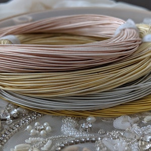 May include: Close-up of four spools of metallic wire. The spools are stacked on top of each other. The wire is in shades of rose gold, gold, silver, and a darker silver. The spools are sitting on a white fabric with pearl-like embellishments.