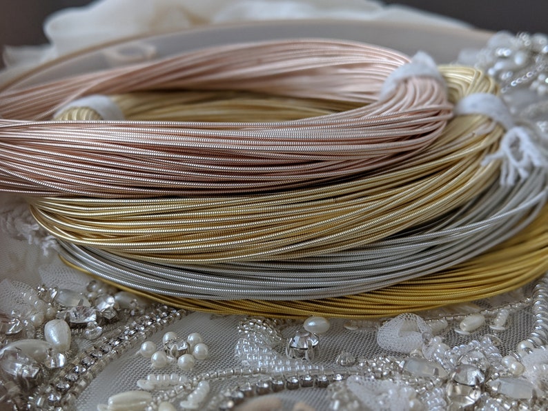 May include: Close-up of four spools of metallic wire. The spools are stacked on top of each other. The wire is in shades of rose gold, gold, silver, and a darker silver. The spools are sitting on a white fabric with pearl-like embellishments.