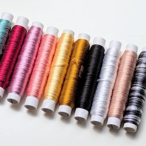 Pode incluir: Uma coleção de dez carretéis cilíndricos de linha de bordar metálica dispostos em uma fileira diagonal. As cores incluem azul-petróleo, magenta, rosa claro, coral, ouro amarelo, ouro escuro, preto, prata, ouro rosa e um padrão listrado em preto e branco.