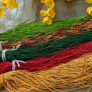 May include: Bundles of colorful elastic cords in green, orange, dark green, red, and gold. The cords are tied with white string. Yellow flowers are in the background.