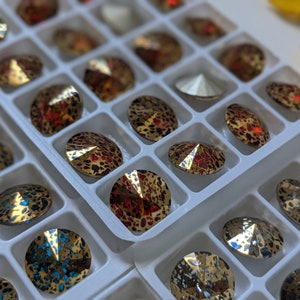 May include: A close-up of a white plastic container filled with various round, iridescent rhinestones. The rhinestones are arranged in rows and have a variety of colors, including gold, red, and blue.