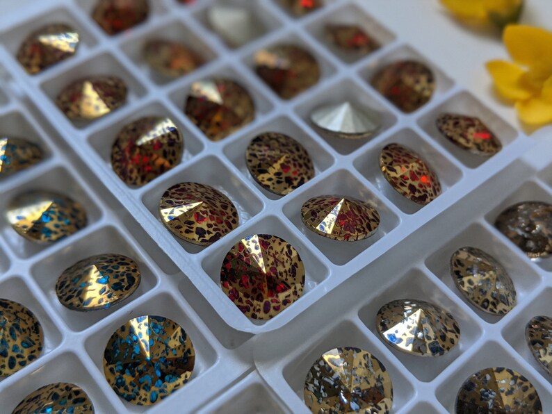 May include: A close-up of a white plastic container filled with various round, iridescent rhinestones. The rhinestones are arranged in rows and have a variety of colors, including gold, red, and blue.