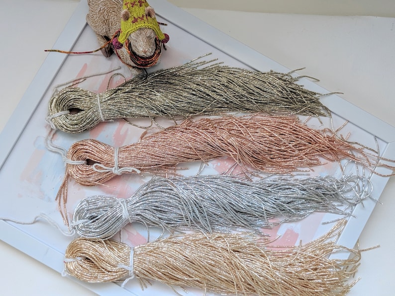 May include: Four spools of metallic thread in gold, silver, rose gold, and green. The thread is arranged in a fan shape on a white surface.