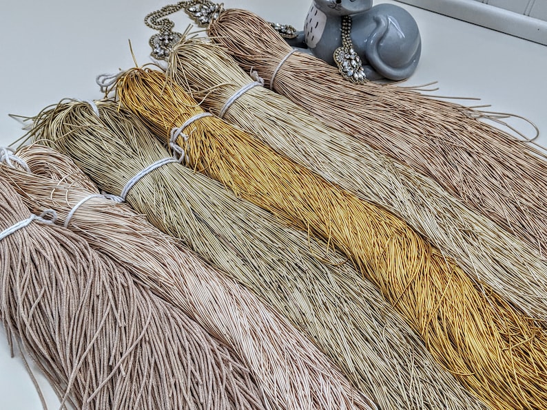 May include: A collection of six spools of metallic thread in various shades of beige, gold, and brown. The thread is arranged in a fan shape, with the ends of the thread extending out from the spools.