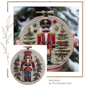 Peut inclure: Broderie sur cerceau représentant un casse-noisette en uniforme rouge et bleu, avec un chapeau noir et une barbe blanche. Le motif comprend des sapins de Noël et des flocons de neige, sur un tissu beige clair. Le texte "NutCracker" est visible.