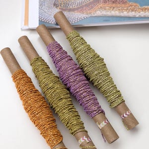 May include: Four cardboard tubes wrapped with metallic glitter thread in orange, gold, purple, and a second gold shade. Each tube is secured with a small piece of patterned adhesive tape at the end, displaying a shimmering texture and vibrant colors.