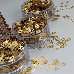 Puede incluir: Tres recipientes transparentes llenos de confeti metálico dorado. Los recipientes están dispuestos en una fila de izquierda a derecha. El confeti del primer recipiente es de forma cuadrada, el confeti del segundo recipiente es de forma redonda y el confeti del tercer recipiente es una mezcla de formas cuadradas y redondas. Hay algo de confeti esparcido sobre la superficie blanca.