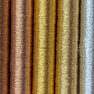 Op de afbeelding: Een verzameling van vijf verticale klossen metallic borduurgaren naast elkaar. De kleuren omvatten donker koper, licht koper, helder goud, gedempt goud en glinsterend zilver, allemaal met een gladde, reflecterende afwerking van de draden.