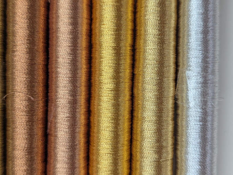 Op de afbeelding: Een verzameling van vijf verticale klossen metallic borduurgaren naast elkaar. De kleuren omvatten donker koper, licht koper, helder goud, gedempt goud en glinsterend zilver, allemaal met een gladde, reflecterende afwerking van de draden.