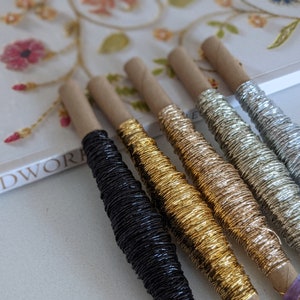 May include: Five spools of metallic embroidery thread in black, gold, copper, silver, and white. The spools are wrapped in brown paper and have a purple label.