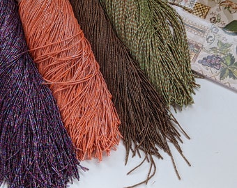 May include: Four spools of seed beads in different colors: purple, orange, brown, and green. The beads are arranged in a row on a white surface.