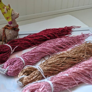 Puede incluir: Cuatro carretes de hilo metálico en tonos rojo, rosa, dorado y rosa claro. El hilo está dispuesto en forma de abanico sobre una superficie blanca.