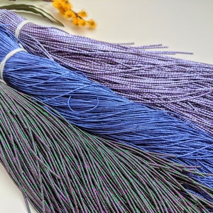 以下が含まれることがあります： 異なる色の伸縮性のあるコードが3つの糸巻きに巻かれています。ピンクがかった光沢のあるグリーン、ブルー、白がかった光沢のあるパープルです。