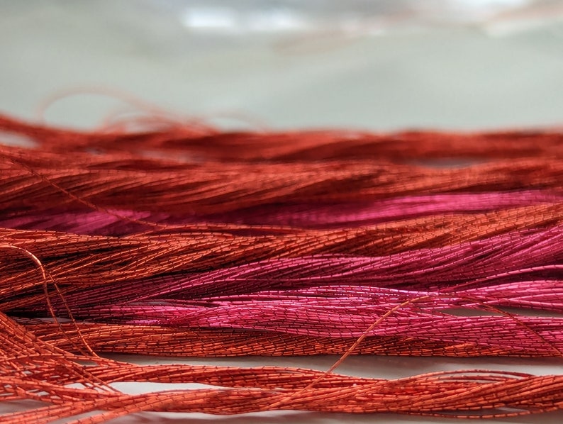 Op de afbeelding: Een verzameling metallic koperkleurige en diep magentakleurige borduurgarens gerangschikt in losse, horizontale strengen. De vezels vertonen een hoogglanzende, reflecterende kwaliteit met een duidelijke gedraaide textuur, wat een levendig contrast cre&euml;ert tussen de warme oranje koperkleuren en de koele, rijke roze-rode tinten.