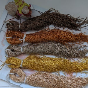 May include: Five spools of metallic thread in various shades of gold, brown, and beige. The thread is arranged in a stack, with the brown spool on top and the beige spool on the bottom.