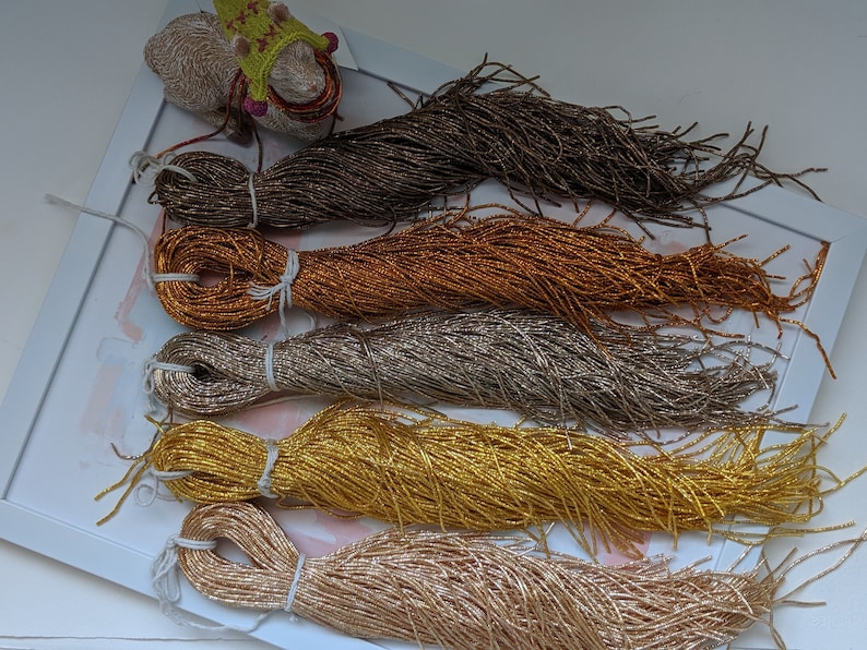 May include: Five spools of metallic thread in various shades of gold, brown, and beige. The thread is arranged in a stack, with the brown spool on top and the beige spool on the bottom.