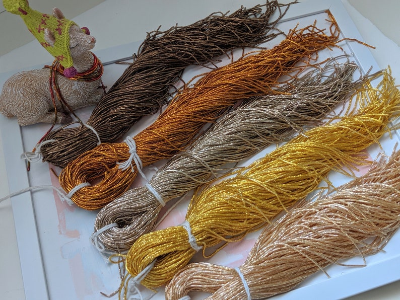 May include: Five spools of metallic thread in various shades of gold, brown, and beige. The thread is arranged in a fan shape on a white surface.