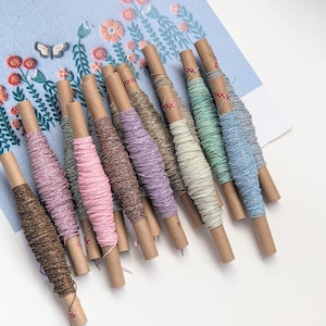 May include: A collection of 12 spools of embroidery floss in various pastel colors, including pink, purple, blue, and green. The spools are wrapped in brown paper and have a red and white striped tape around them.