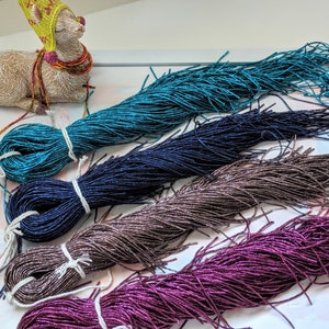 May include: Four spools of metallic thread in teal, navy blue, brown, and purple. The thread is wound around white spools and is arranged in a row on a white surface.