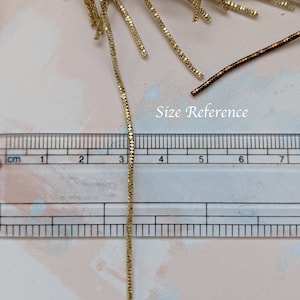 May include: A gold chain measuring 5 cm in length, next to a ruler for size reference.  The chain is made of small, linked squares.
