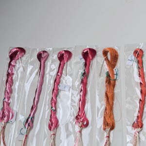 Op de afbeelding: Zes individuele bundels borduurgaren, elk verpakt in een doorzichtige plastic hoes. De collectie bevat vier tinten roze en magenta, &eacute;&eacute;n oranje en &eacute;&eacute;n roodachtig oranje, allemaal voorzien van kleine witte papieren labels aan de draad.