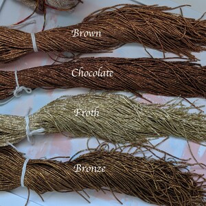 May include: Four spools of metallic seed beads in different colors: brown, chocolate, froth, and bronze. The beads are arranged in rows on a white surface.