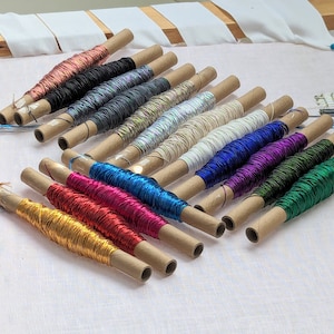 Könnte beinhalten: Eine Sammlung von 16 Spulen mit Metallic-Garn, jede in braunes Papier gewickelt und in einer Reihe angeordnet. Die Fäden sind in verschiedenen Farben, darunter Gold, Silber, Rot, Blau, Grün und Lila.