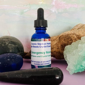May include: A blue glass bottle with a dropper top, labeled "Cosmic Stardust Healing Light Beads Crystal Essence Emergency Rescue Flame and Crystal Essence" and "formulas of rare and not crystals and minerals". The bottle is sitting on a purple surface with a green crystal, a blue glass orb, and a black stone.