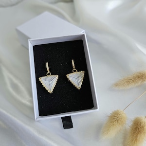 Op de afbeelding: Een paar gouden driehoekige oorbellen met witte pareldetails, gepresenteerd in een witte doos met een zwarte binnenkant.