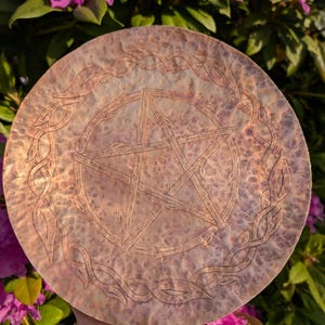 May include: A copper-colored, round metal plate with an etched pentagram in the center, surrounded by a decorative vine-like border. The plate has a textured surface and is held in front of a background of pink flowers and green foliage.