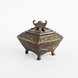 May include: A bronze-colored, four-legged incense burner with a decorative lid. The lid features an ornate handle and cut-out designs. The body of the burner has etched patterns and a geometric shape, suggesting an antique or vintage style.