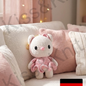 Könnte beinhalten: Eine handgefertigte Häkelkatzenpuppe in Weiß und Rosa mit einer rosa Schleife und einem Kleid. Die Puppe hat schwarze Augen und eine rosa Nase. Sie sitzt auf einem weißen Sofa mit rosa Kissen. Eine deutsche Flagge ist im Bild.