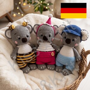 Könnte beinhalten: Drei handgefertigte Koala-Bären-Puppen aus Häkelarbeit in einem Weidenkorb. Die Koalas sind grau mit verschiedenen Outfits: einem Bienenkostüm, einem rosa Outfit mit Hut und einem blauen Outfit mit Mütze. Der Korb steht auf einer flauschigen weißen Oberfläche.
