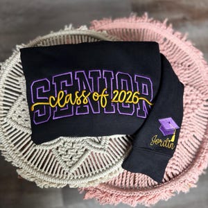 May include: Black sweatshirt with embroidered text "SENIOR class of 2026" in purple and gold. The sleeve features a graduation cap and the name "Jordin" in gold. The sweatshirt is displayed on a macrame and pink fringed surface.